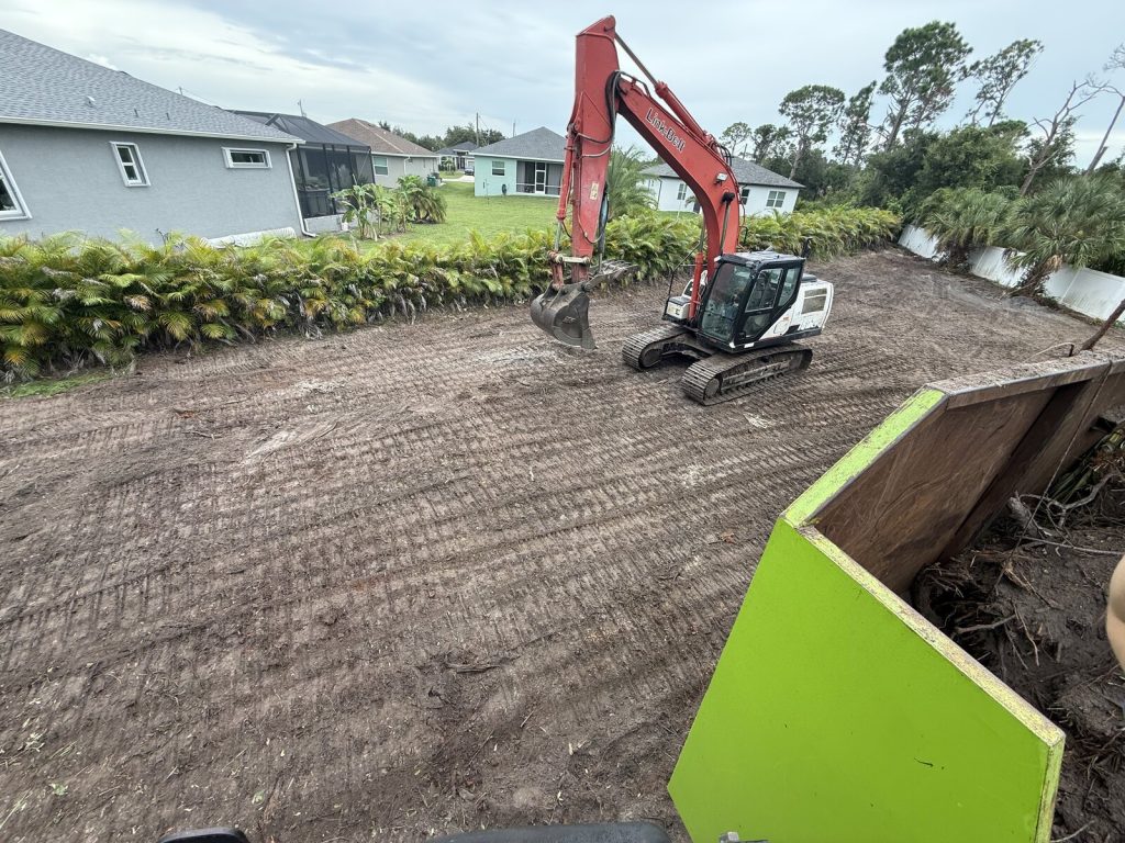 Tracked excavator grading a residential lot beside established palms and fencing