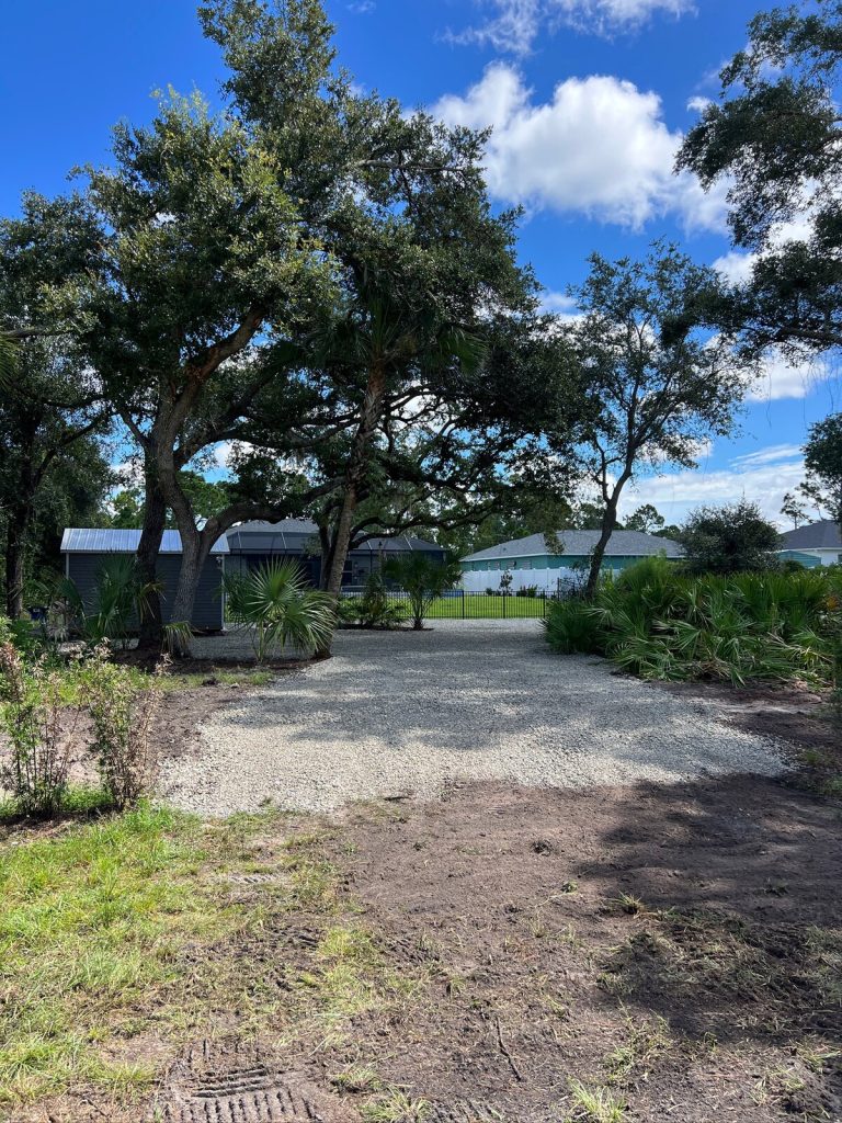 Gravel driveway leading past mature trees and palms toward a backyard shed