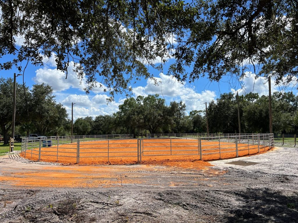 Wide view of a new horse arena with red clay footing and steel fencing